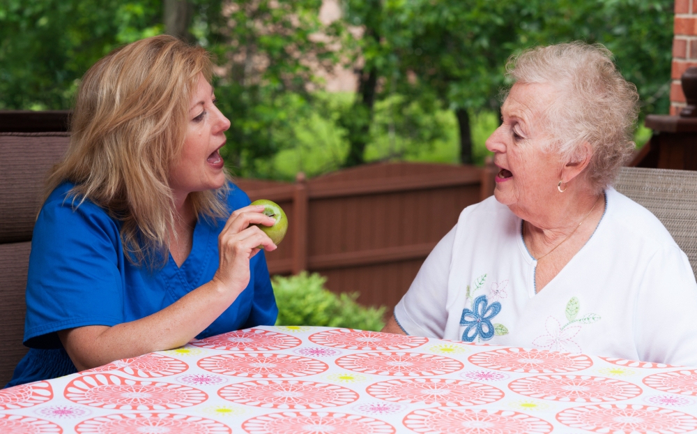 A speech therapist incorporating an apple into a speech therapy session with an elderly stroke patient, utilizing it as a tool for speech exercises and oral motor skill development, fostering communication and rehabilitation.